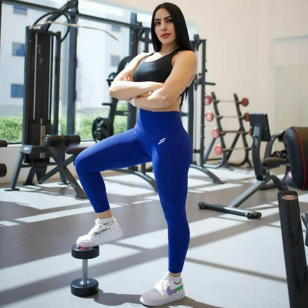 A woman in a gym stands confidently with arms crossed. She wears a black tank top and blue leggings, resting one foot on a dumbbell. Behind her are various gym equipment like weights and exercise machines. Sunlight streams in through large windows, casting shadows on the floor.