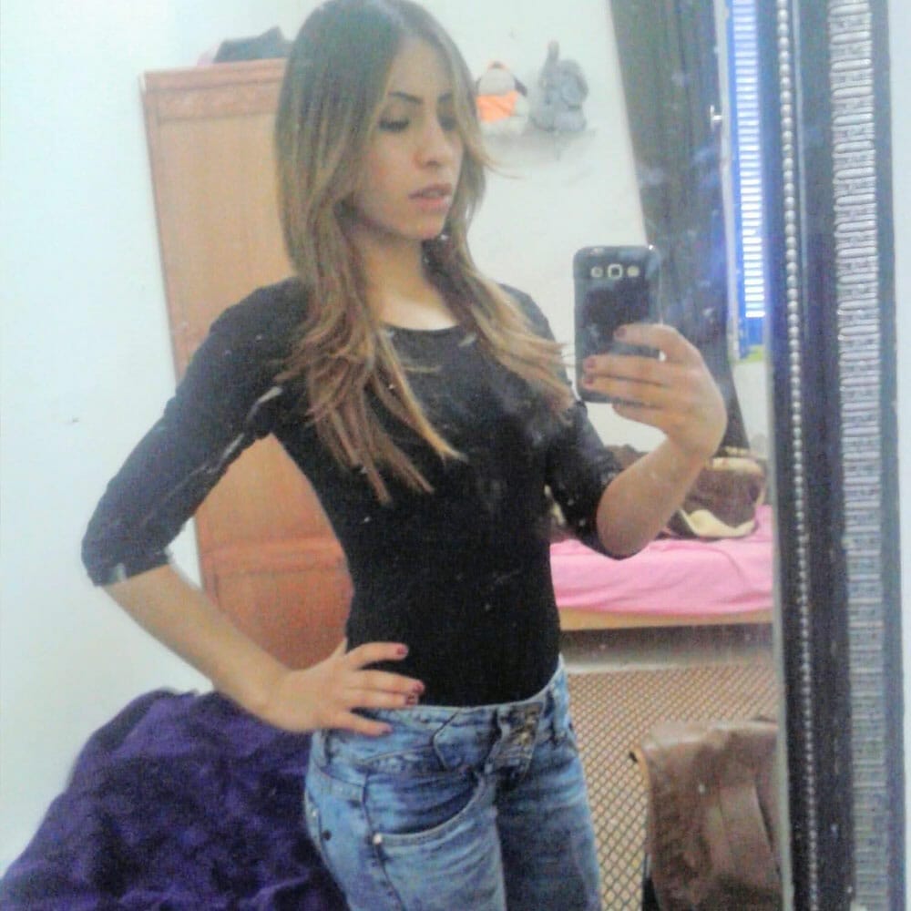 A person wearing a black top and jeans takes a mirror selfie in a room. They have long, light brown hair. In the background, there's a wooden wardrobe, a bed with a pink blanket, and some items hanging on the wall. The mirror has some smudges.